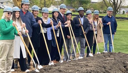 Current and past members of the Library Board of Trustees and the Executive Director of the Library wearing hard hats and holding shovels behind a mound of dirt at the site of the future library on Maple Avenue in Roselle, Illinois.
